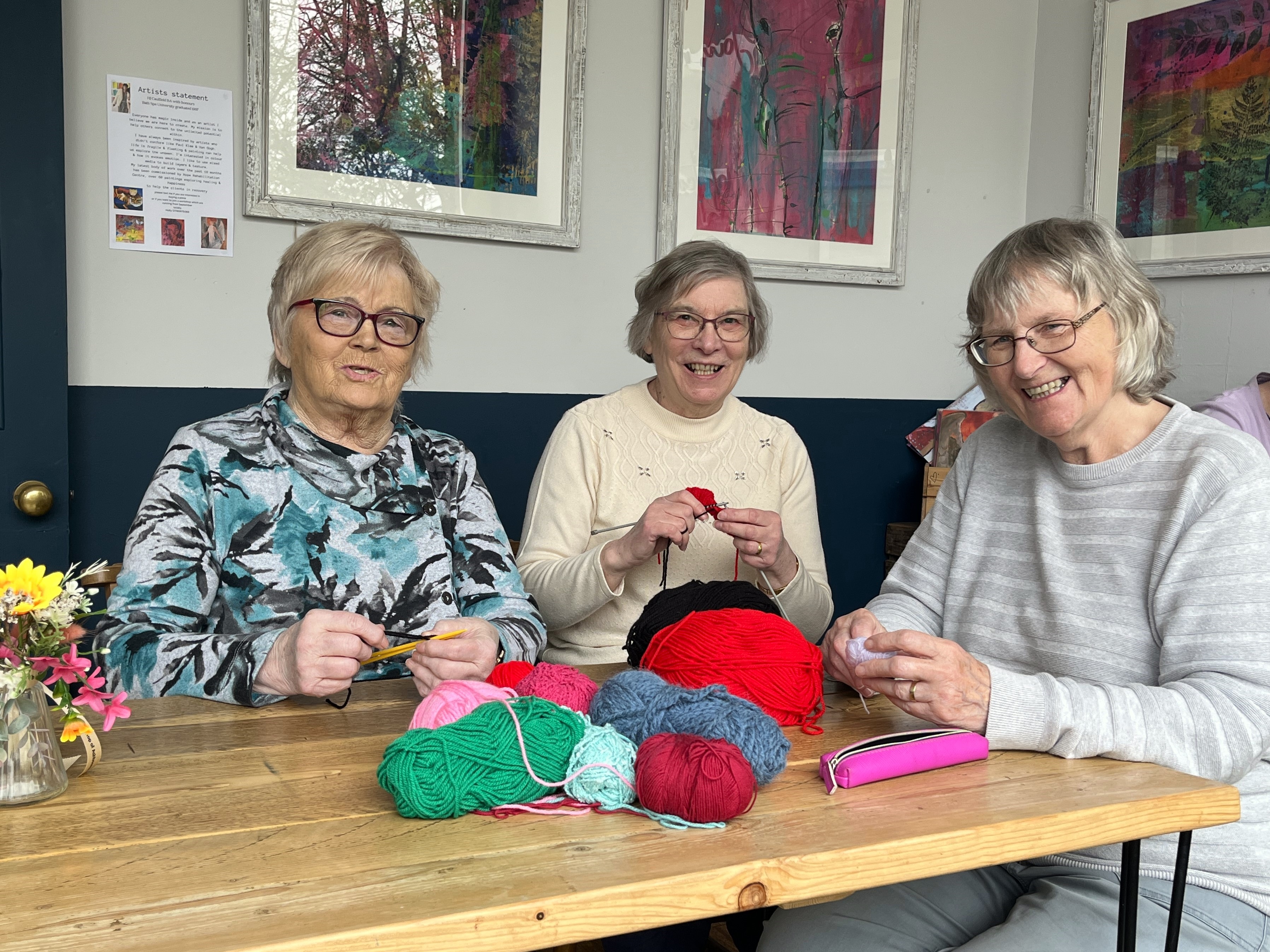 Three smiling people sat holding knitting at a table covered with balls wool 