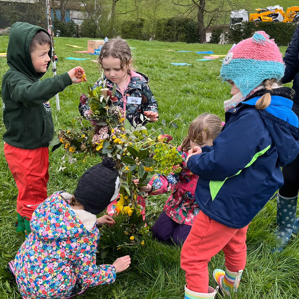 Photo of children decorating a cross shape with flowers. The children are wearing woolly hats and wellies.